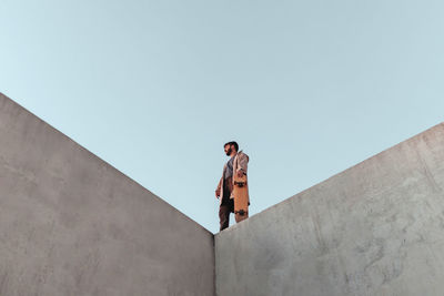 From below of young bearded male skater standing on high concrete ramp in skatepark with skateboard looking away