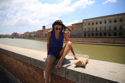 Smiling young woman sitting on retaining wall by river in city