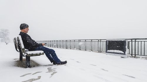 Man sitting on snow covered railing against clear sky