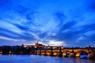 Illuminated bridge over river by buildings against sky at dusk