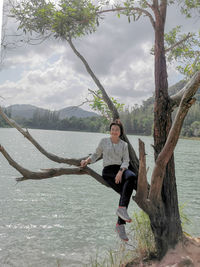 Full length of man sitting on tree by lake against sky