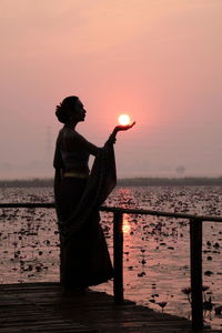 Optical illusion of woman holding sun while standing by lake against sky during sunset