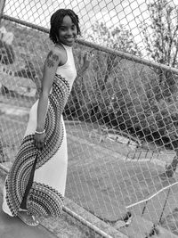 Portrait of smiling young woman wearing dress while standing by chainlink fence