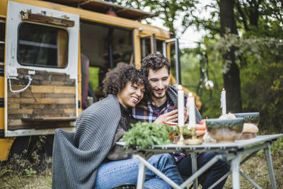 Smiling young couple sharing mobile phone while wrapped in blanket together during camping in forest