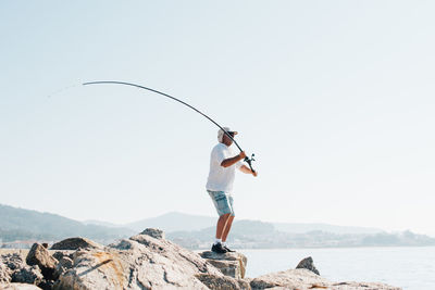 Side view of enthusiastic man in cap fishing and throwing rod into sea water standing on stony shore in bright day