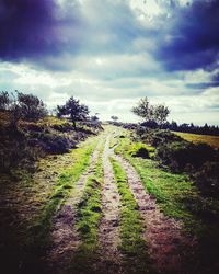 Dirt road leading towards trees against cloudy sky