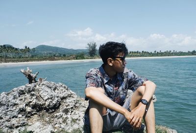 Young man sitting on rock by sea against sky