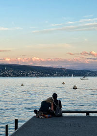 Rear view of man sitting on pier over sea against sky during sunset