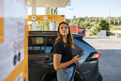 Woman holding smart phone and looking away while refueling car at gas station