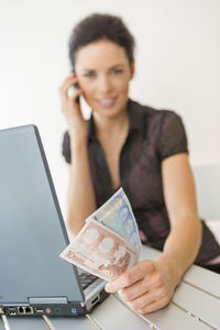 Portrait of smiling businesswoman with currency using phone by laptop on desk