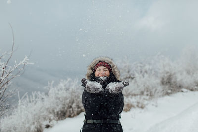 Cheerful woman in warm clothing throwing snow while standing on mountain