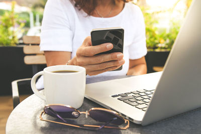 Midsection of woman using phone by coffee cup and laptop on table