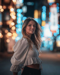 Portrait of smiling young woman standing on illuminated street at night