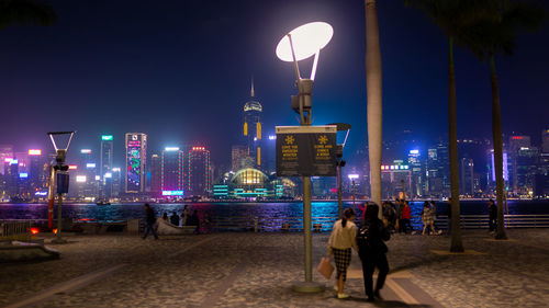 People on footpath against sea and illuminated buildings in city at night