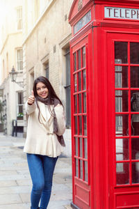 Portrait of smiling young woman gesturing thumbs up while standing by telephone booth in city