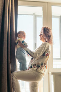 Side view of mother carrying son while standing by window at home