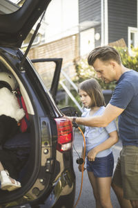 Portrait of mother, father and two daughters standing by car at electric vehicle charging station