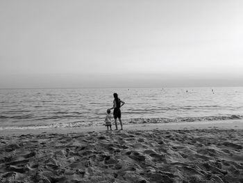 Rear view of mother standing with daughter on shore at beach against clear sky
