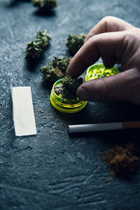 From above faceless man grinding dry cannabis bud while rolling blunt on grunge surface at home
