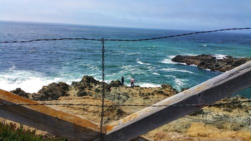 Disruptive view through wire fence of couple standing on ocean cliff. 