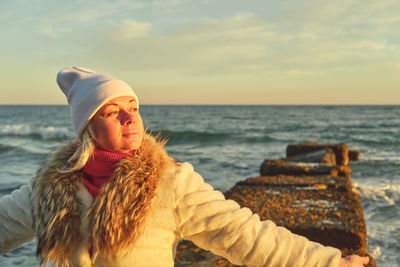 A woman with blond hair in a hat and a fur coat walks along the sea along a stone breakwater