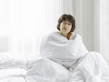 Sleepy woman is sitting in bed, completely covered with white blanket. it's hard to wake up