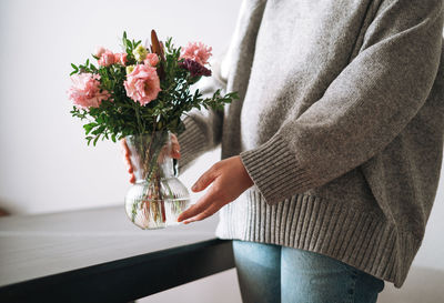 Young woman in grey knitted sweater holding vase with bouqet of flowers at home