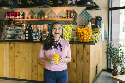 Cheerful mature woman holding mason jar with fresh juice while walking in cafe