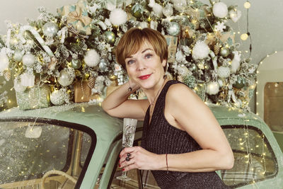 An adult woman drinks champagne at a vintage car next to a christmas tree on new year's eve