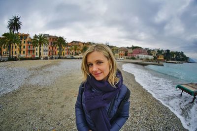 Portrait of smiling young woman standing at beach against cloudy sky