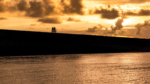 Silhouette men cycling against cloudy sky during sunset