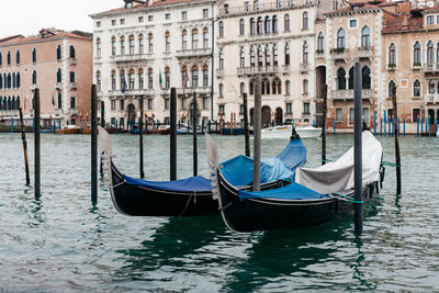 Covered gondolas moored by wooden posts in canal against buildings