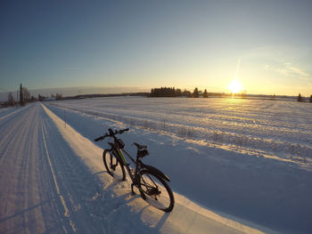 Man riding bicycle on snow covered landscape against sky during sunset