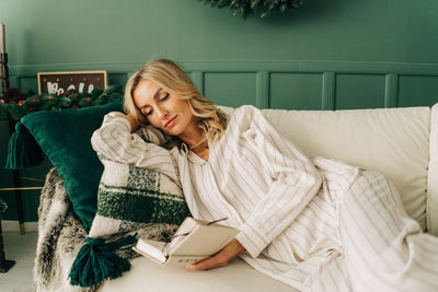 A woman in pajamas is lying on a cozy sofa in a living room decorated for christmas reading a book.