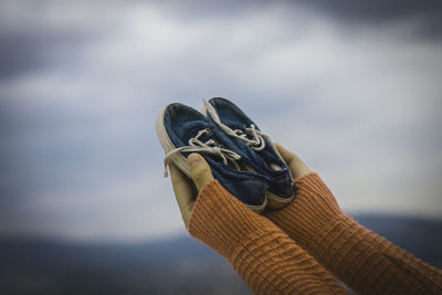 Cropped hands holding shoes against sky