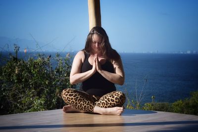 Woman meditating while sitting on wooden floor against sea and sky