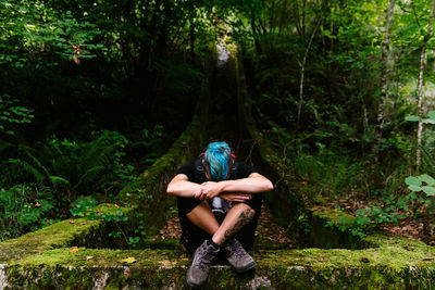 Unrecognizable traveling woman resting on stone mossy border in green woods while embracing knees during trekking