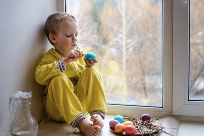 Full length of cute boy holding easter egg while sitting on window sill at home