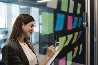 Female entrepreneur planning strategies while standing near glass wall with adhesive note at office