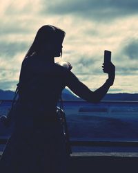 Mother with son taking selfie while standing by railing against cloudy sky