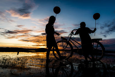 Silhouette children with bicycle standing by lake against sky during sunset