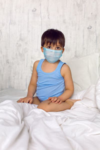 Boy in a blue t-shirt and a medical mask sits on a bed in quarantine during the covid-19 epidemic