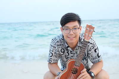 Portrait of young man holding violin while sitting at beach against sky during sunset