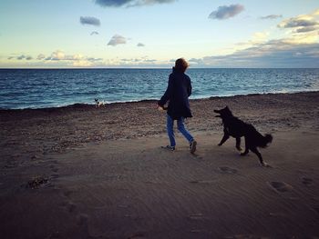 Man and dog standing on beach against sky during sunset