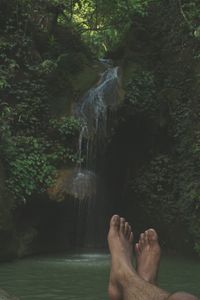 Low section of man relaxing by waterfall in forest