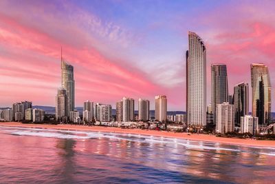 View of modern buildings by sea against sky during sunset