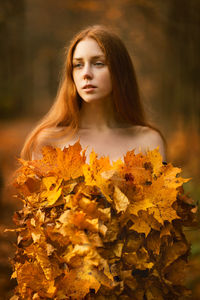 Thoughtful young woman covered with leaves standing at forest during autumn