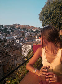 Young woman sitting with drink while looking at city against clear sky