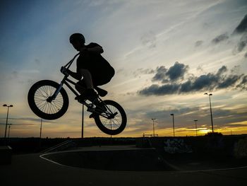 Silhouette man riding bicycle in mid-air against cloudy sky at sunset