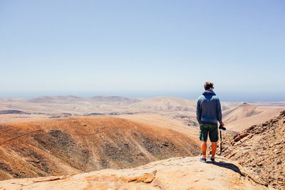 Rear view of man standing on rock looking at landscape against blue sky